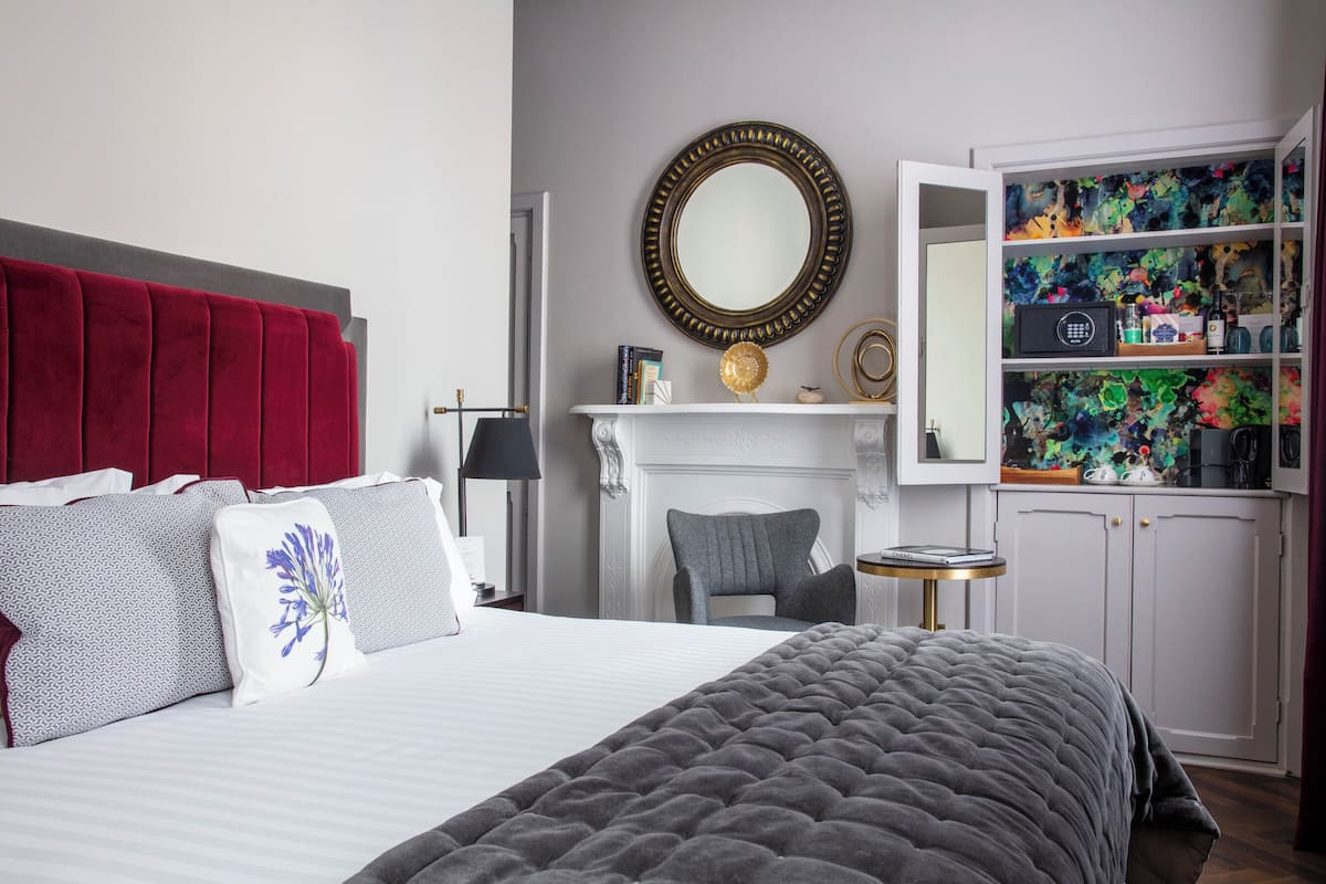 Un dormitorio con cama, cabecera roja, chimenea, espejo y estantería.