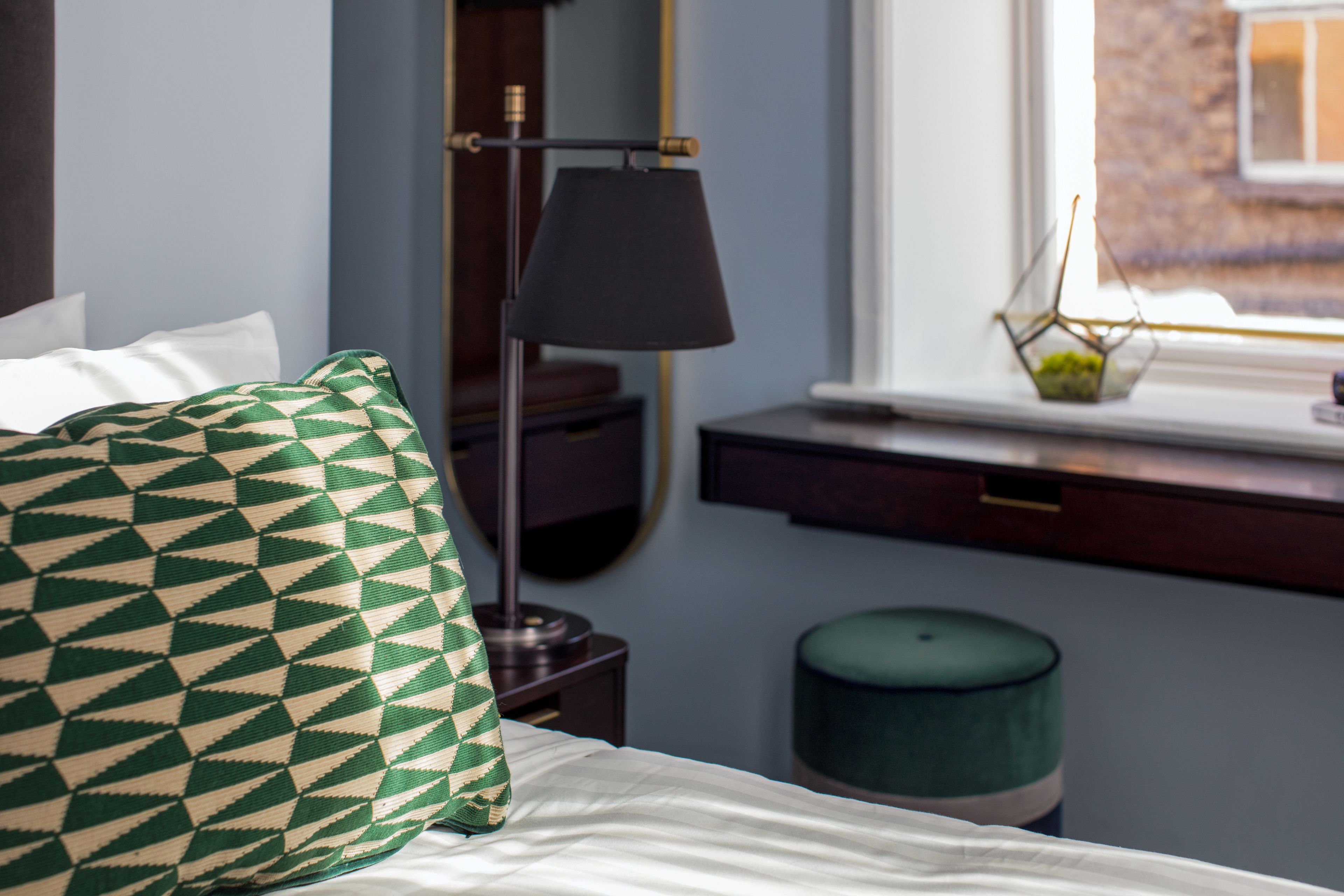 A neatly made bed with a patterned pillow, a bedside lamp, a small table with a vase, and a window with a view of a building.