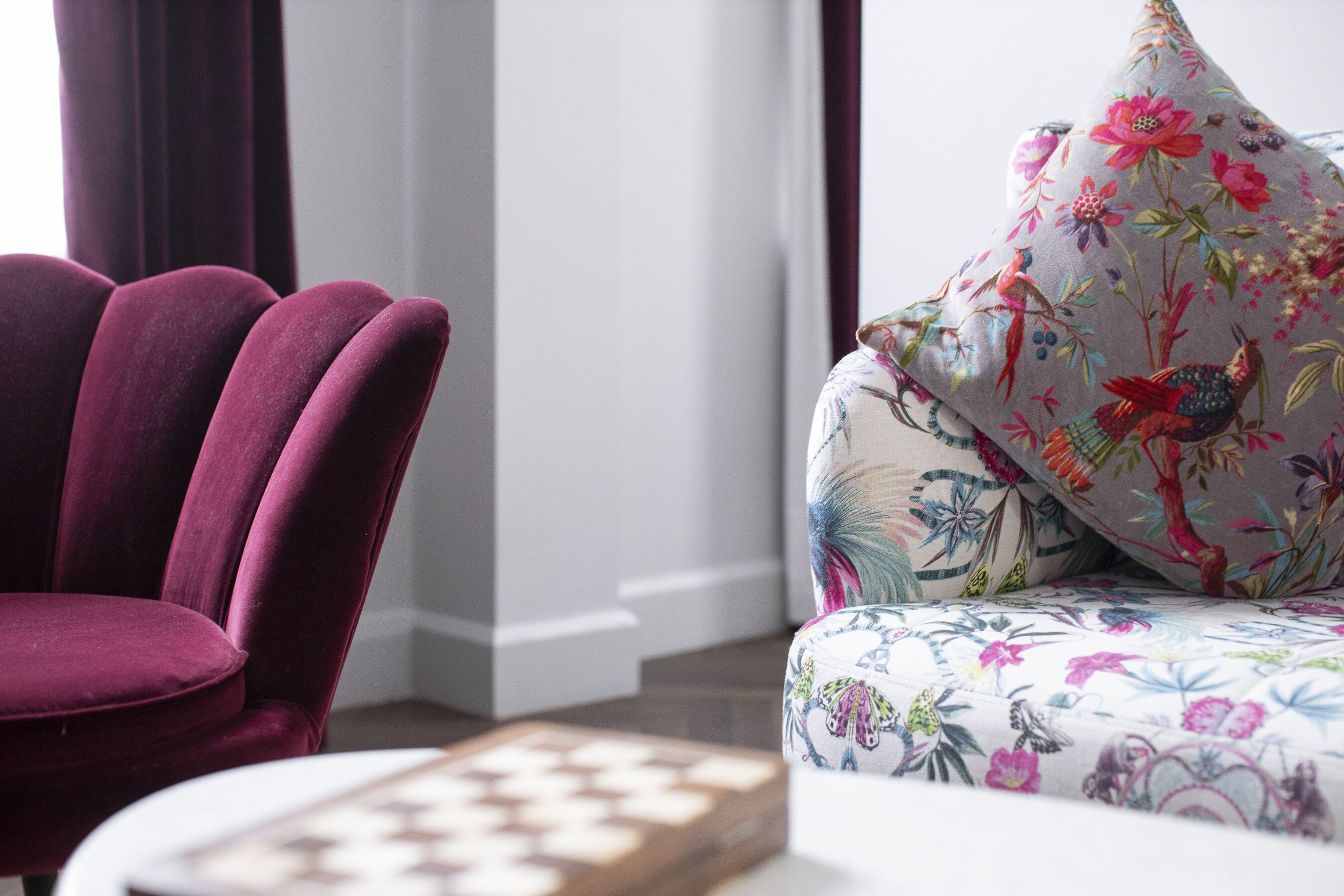 A plush purple armchair and a floral-patterned sofa in a well-lit room with a chessboard on a table.