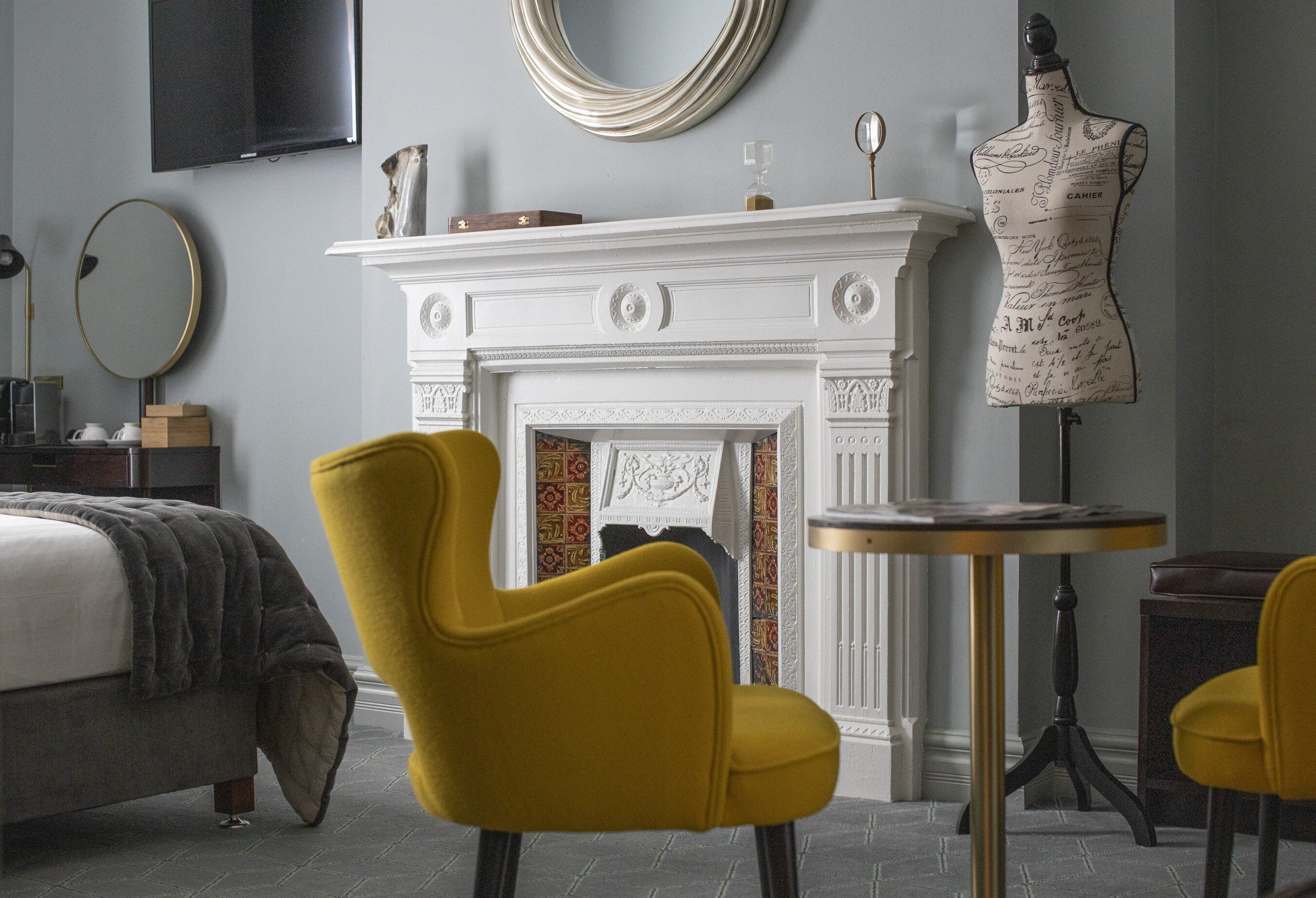 Un dormitorio con una cama, un sillón amarillo, una mesa redonda, una chimenea y un maniquí.