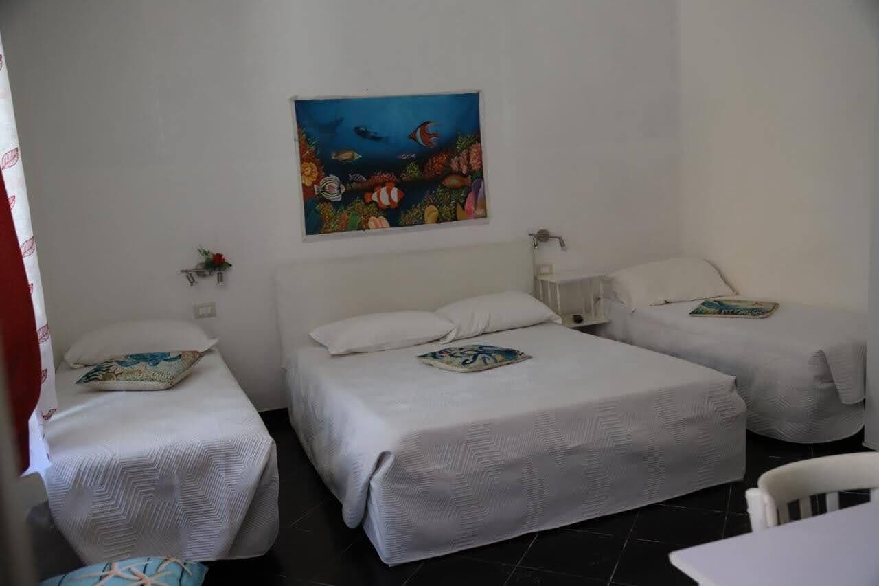 A room with two single beds, a painting of an underwater scene, a small shelf with a flower, and a white chair.