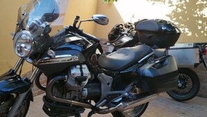 Una motocicleta estacionada sobre una superficie de baldosas, con una pared amarilla y plantas en macetas al fondo.