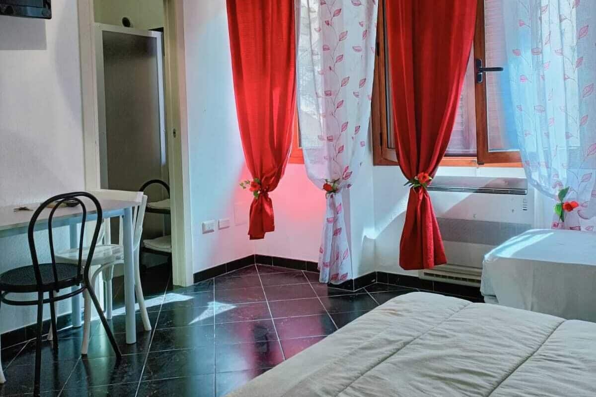 Una habitación de hotel con cama, una mesa de comedor con sillas y una ventana con cortinas rojas.