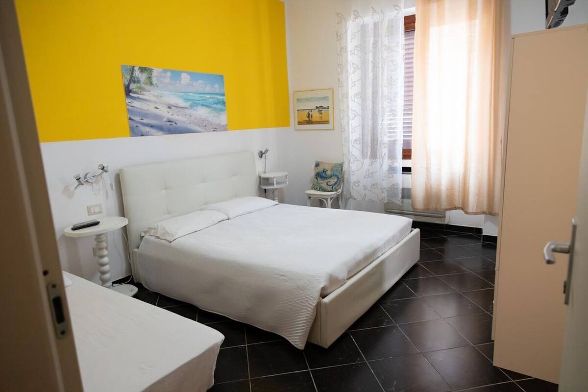Un dormitorio con cama, mesita de noche, una pequeña mesa y una silla. Hay una ventana con cortinas y un cuadro en la pared.