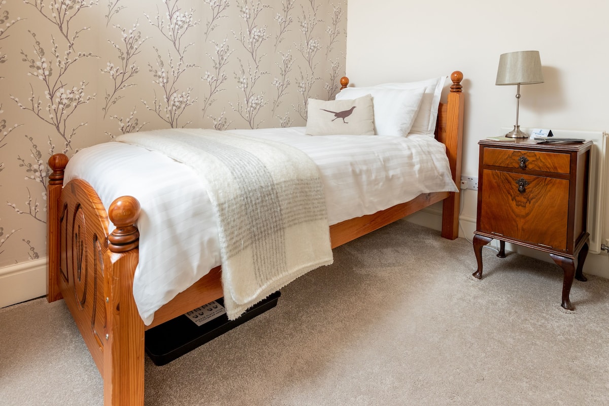 Una cama bien hecha con cabecera de madera, una mesita de noche con lámpara, y una pared con papel tapiz floral.