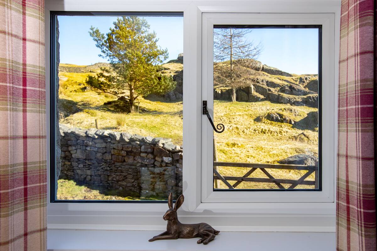 Una ventana con vista a un muro de piedra y una valla de madera, una escultura de metal de un conejo y un cortinaje escocés.