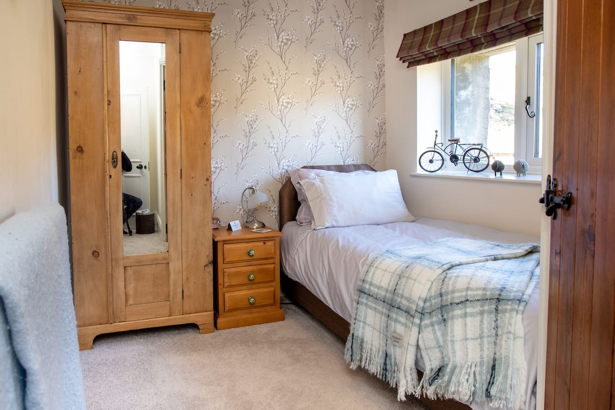 Un dormitorio con un armario de madera, una mesita de noche con lámpara, una cama con manta de cuadros y una ventana con una bicicleta afuera.
