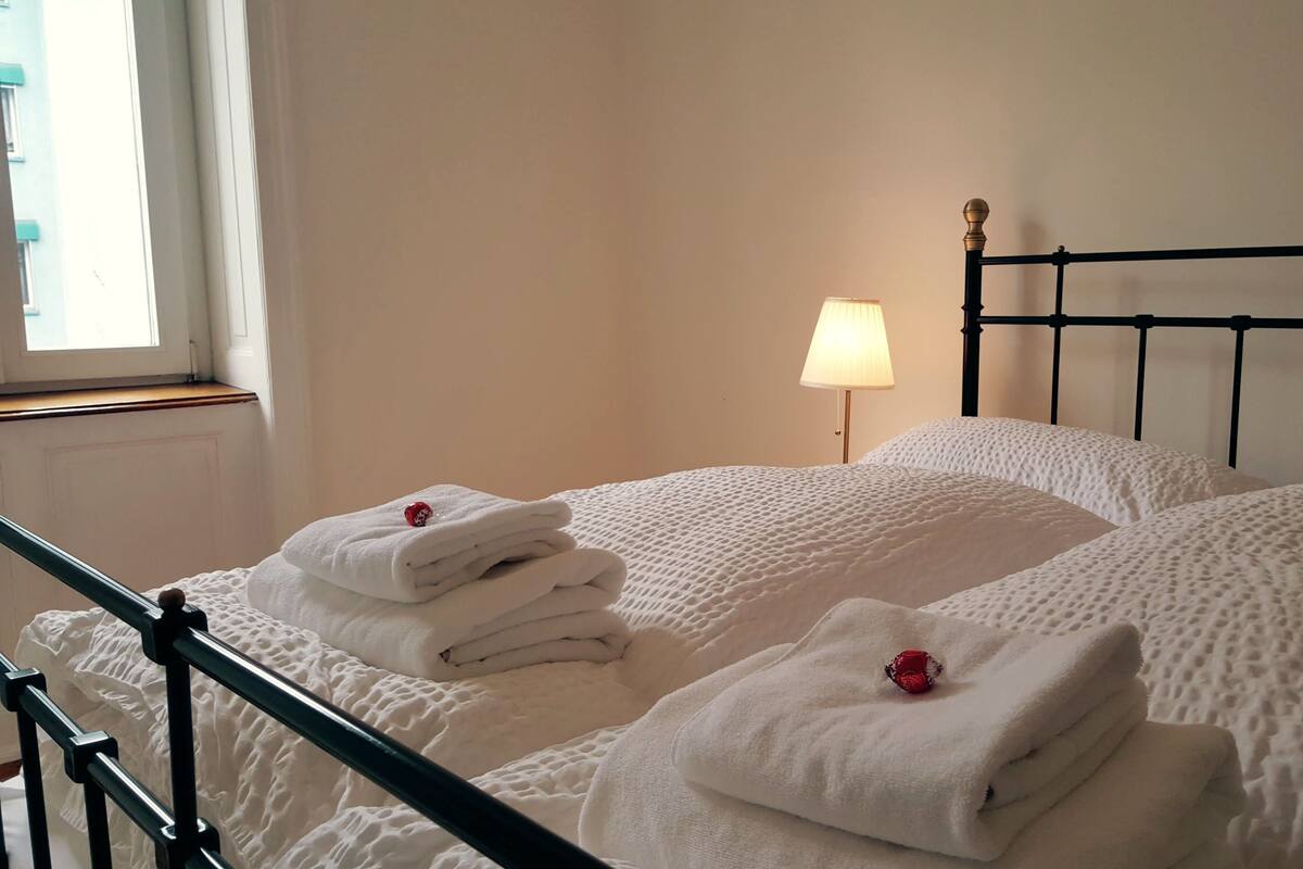 A neatly made bed with white linens and pillows, a bedside lamp, and a window with a view of the outside.