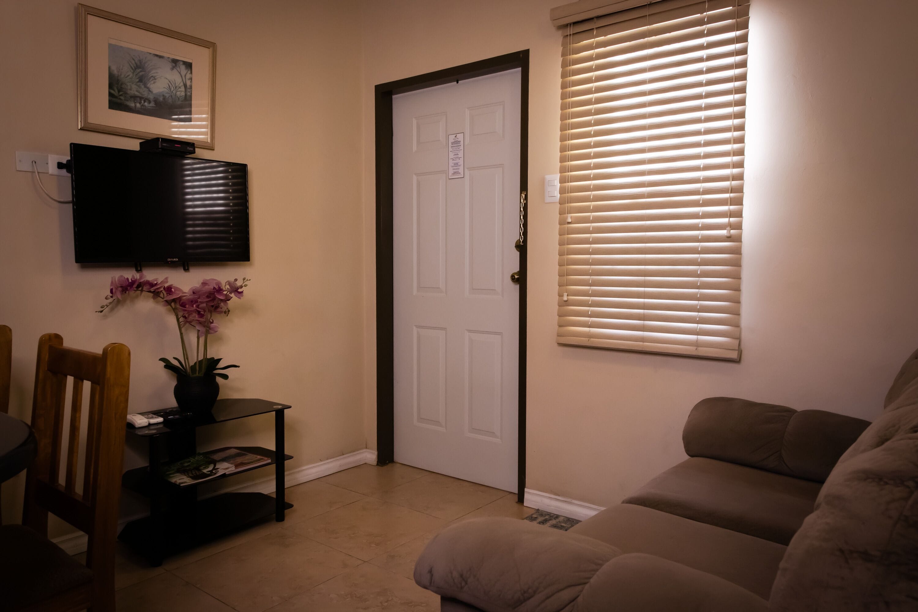 A living room with a beige couch, a TV mounted on the wall, a door with a sign, and a window with blinds.