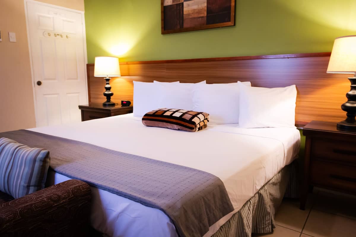 A neatly made bed with white linens, a patterned pillow, and a gray blanket. There are two bedside tables with lamps, a wooden headboard, and a green wall.