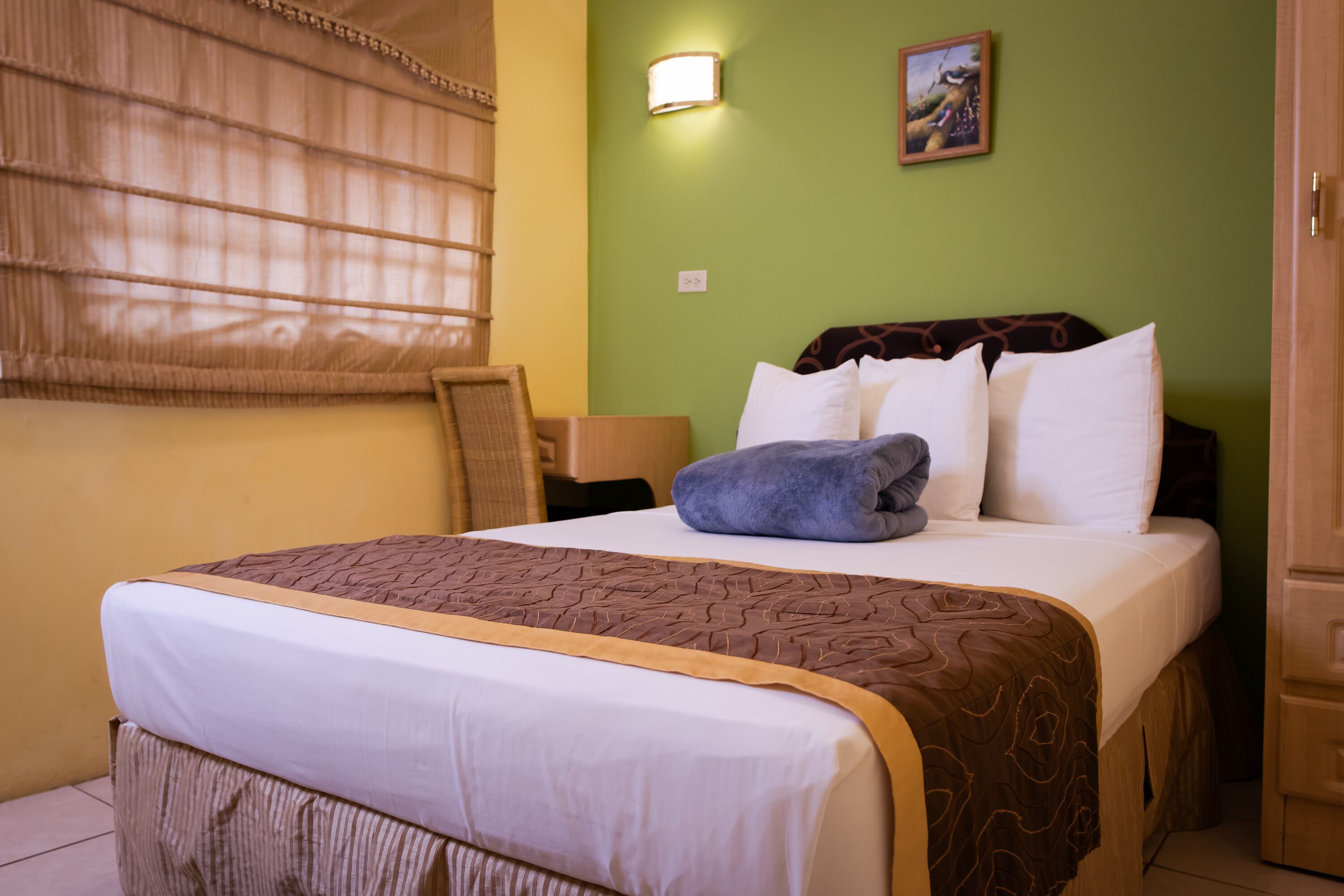 A neatly made bed with a brown and white patterned bedspread, a blue towel, and white pillows. There is a wall-mounted lamp and a painting on the green wall.