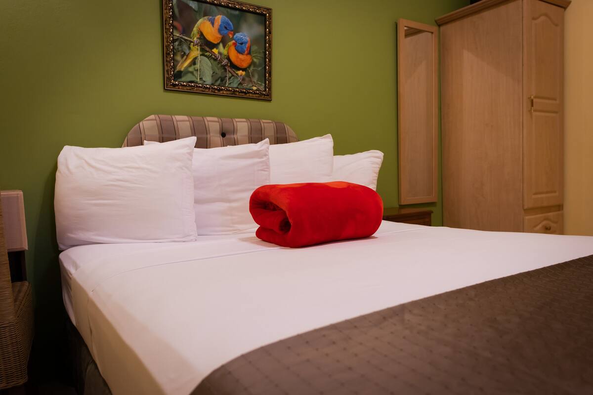 A neatly made bed with white linens and a red blanket, a framed bird painting on the wall, and a wooden wardrobe.