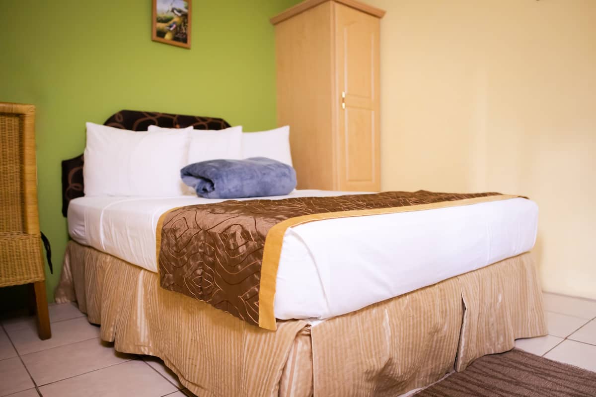 A neatly made bed with a brown and white bedspread, a blue towel, and a wooden wardrobe in a room with green walls and a tiled floor.