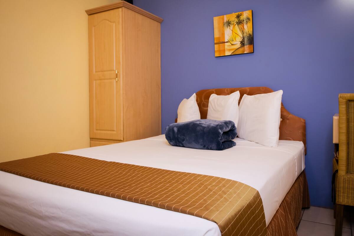 A neatly made bed with a brown striped bedspread, a wooden wardrobe, and a painting of palm trees on the wall.