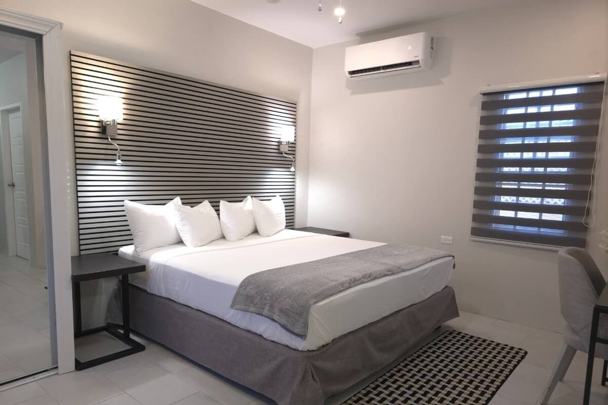 A modern bedroom with a bed, bedside tables, a headboard, an air conditioning unit, a window with blinds, and a chair.