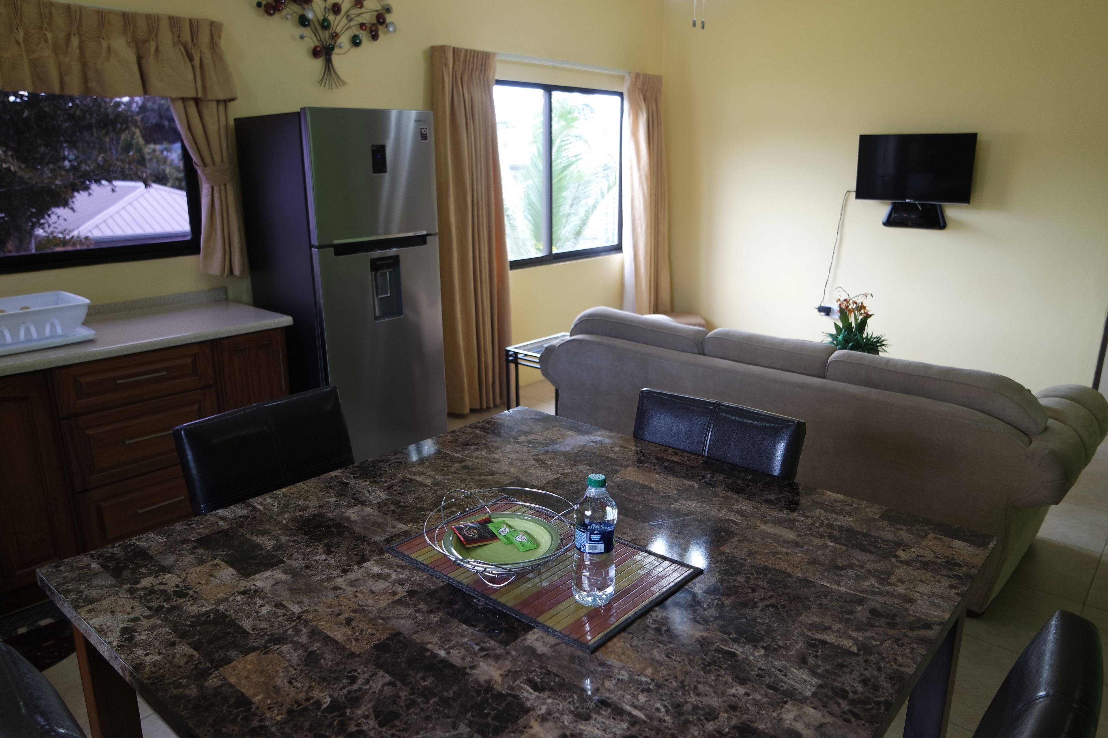 A dining area with a granite table, a water bottle, and a tray with food. A refrigerator and a television are visible in the background.