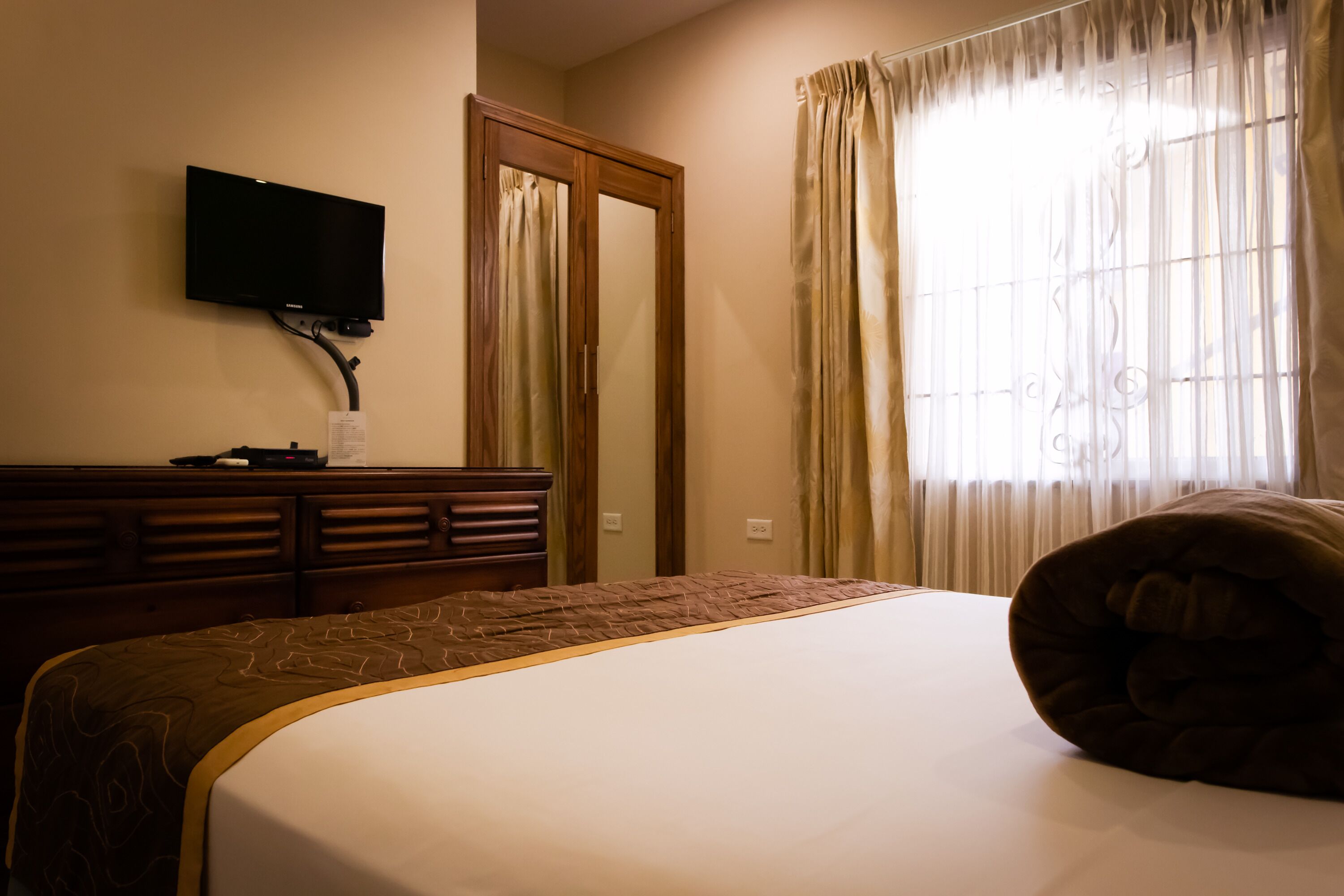 A hotel room with a bed, a television mounted on the wall, a wooden dresser, and a window with curtains.