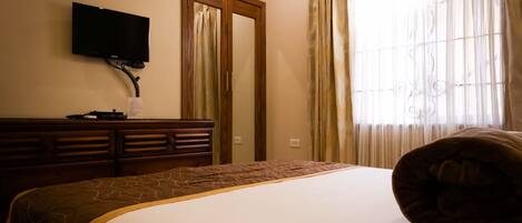 A hotel room with a bed, a television mounted on the wall, a wooden dresser, and a window with curtains.