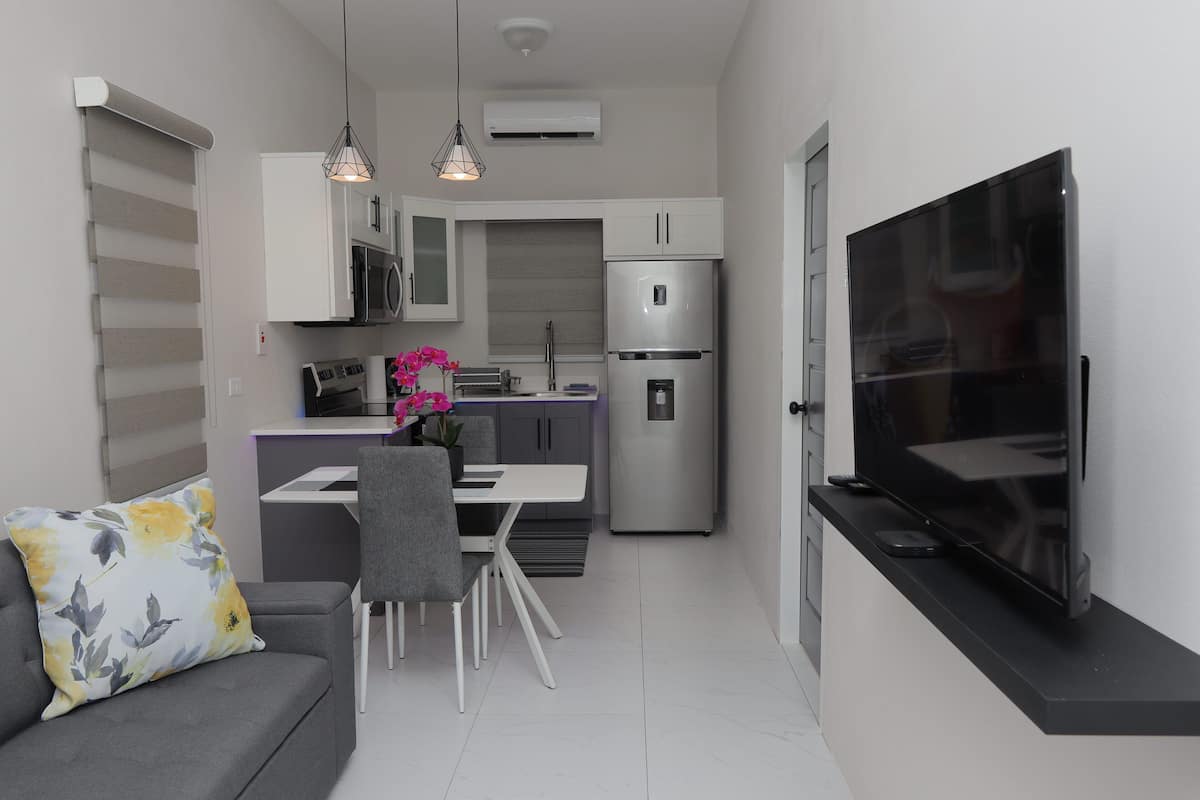 A modern kitchen with white cabinets, a stainless steel refrigerator, a dining area with a table and chairs, and a flat-screen TV mounted on the wall.