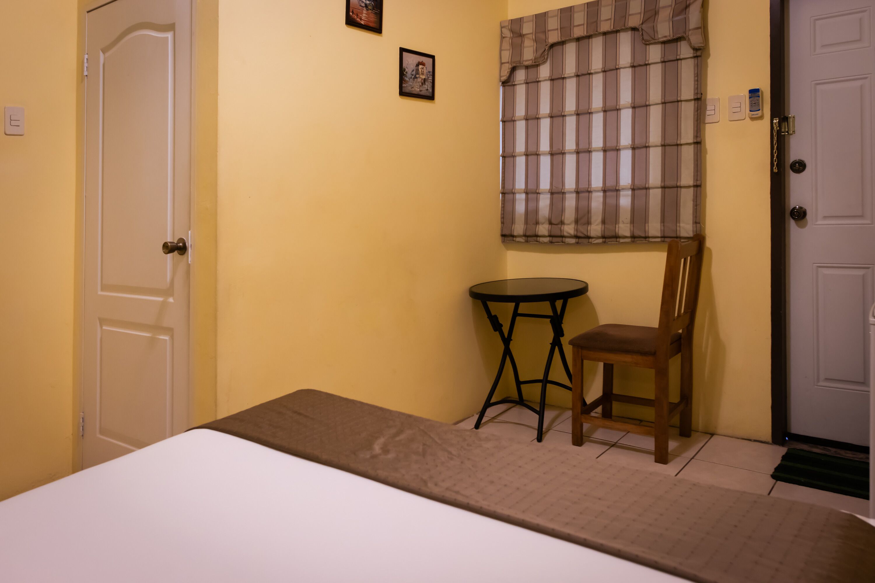 A room with a bed, a small round table, a chair, a window with plaid curtains, and a door.