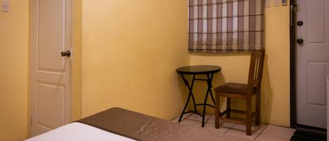 A room with a bed, a small round table, a chair, a window with plaid curtains, and a door.