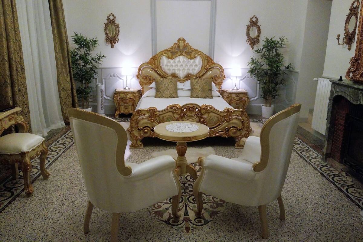 Un dormitorio lujoso con una cama ornamentada, dos sillas, una mesita de noche y un espejo.