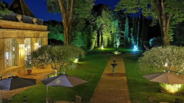 A nighttime garden with a paved walkway, outdoor seating, and illuminated trees. - Château Rieutort (Saint-Pargoire)