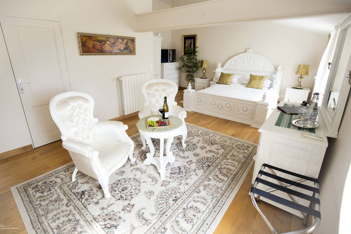 Un dormitorio con una cama grande, una zona de estar con una mesa y una silla. Hay un radiador, una planta y un espejo en la pared.