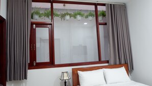 A hotel room with a bed, a nightstand with a lamp, a window with curtains, and hanging plants. - White Lion House (Da Nang)