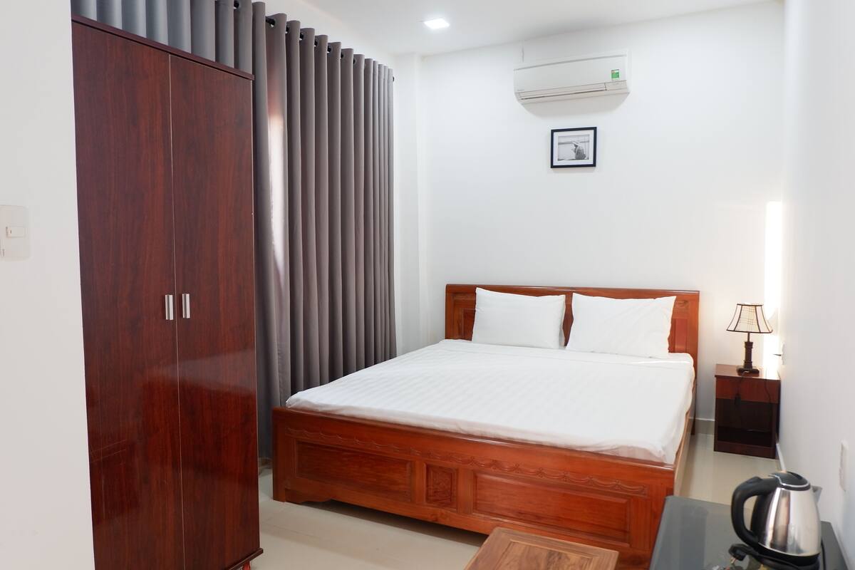 A hotel room with a wooden bed, a wardrobe, a grey curtain, an air conditioning unit, a framed picture, a bedside table with a lamp, and a kettle.