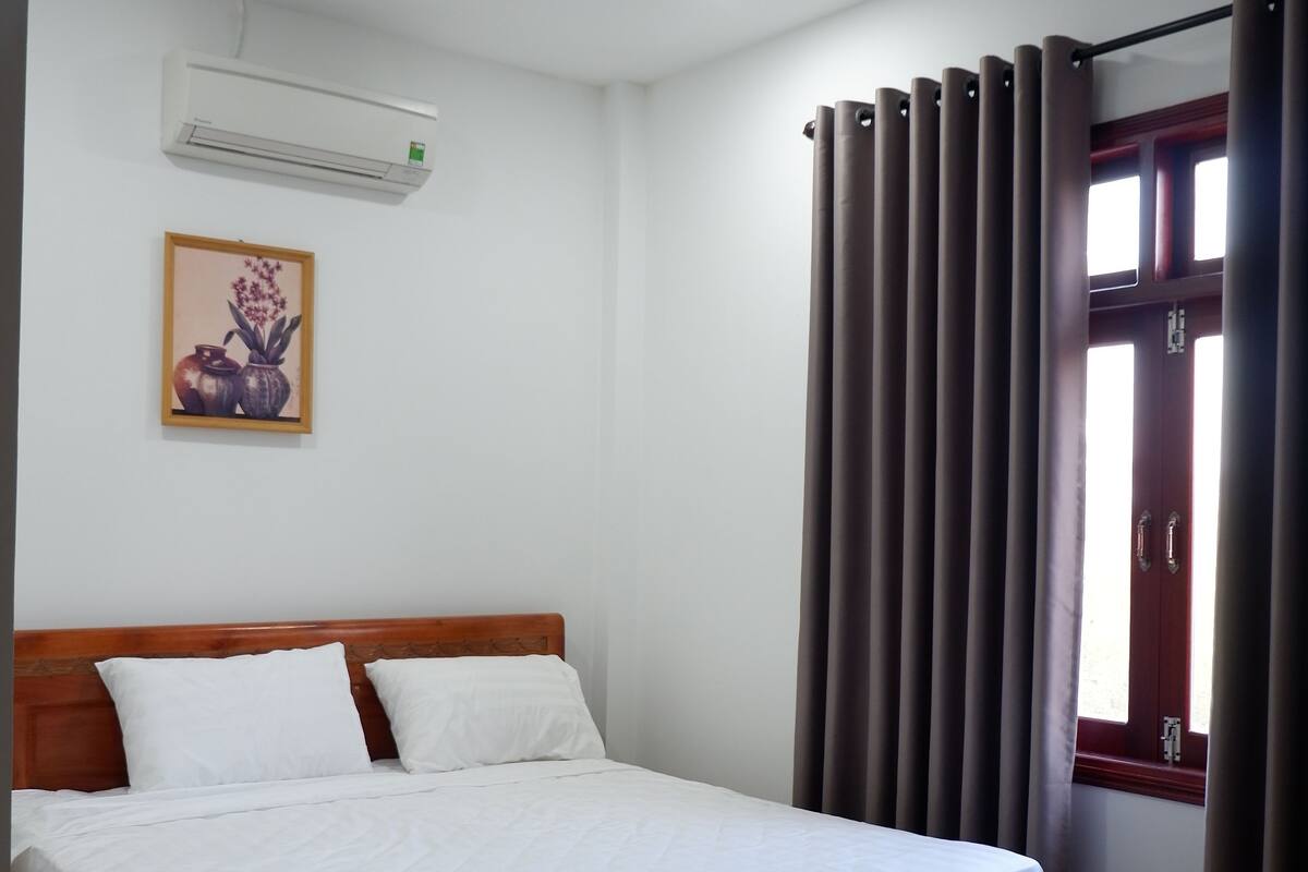 A neatly made bed in a room with a wooden headboard, an air conditioning unit on the wall, and a window with curtains.