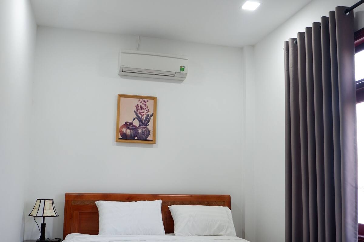 A bedroom with a wooden bed, white bedding, a nightstand with a lamp, an air conditioning unit, and a framed picture on the wall.