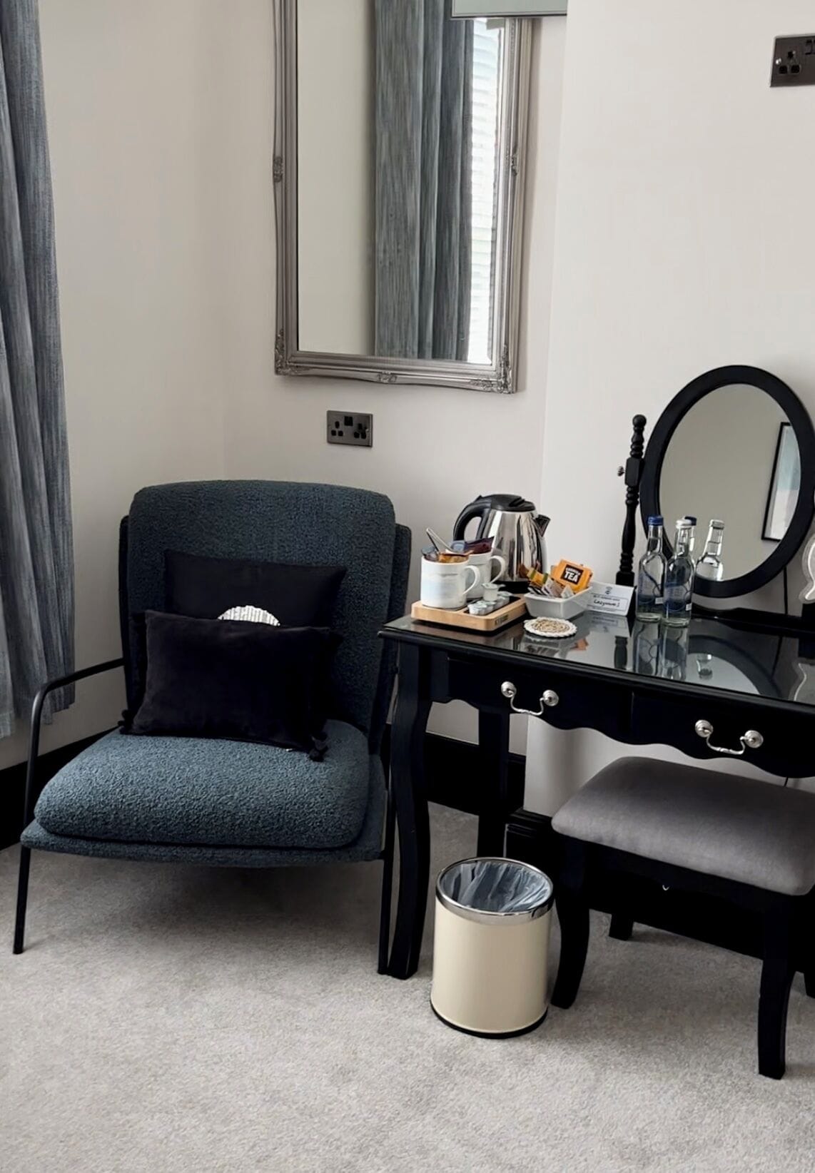 A room with a blue armchair, a black desk with a mirror, a small trash can, and a window with curtains.
