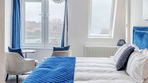 Ein Hotelzimmer mit Bett, Sessel, einem kleinen Tisch, einer Kristallleuchtre und Blick auf eine Stadtkulisse durch große Fenster.