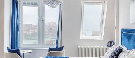 Habitación de hotel con cama, una silla, una mesita, una araña y vistas de la ciudad a través de grandes ventanales.
