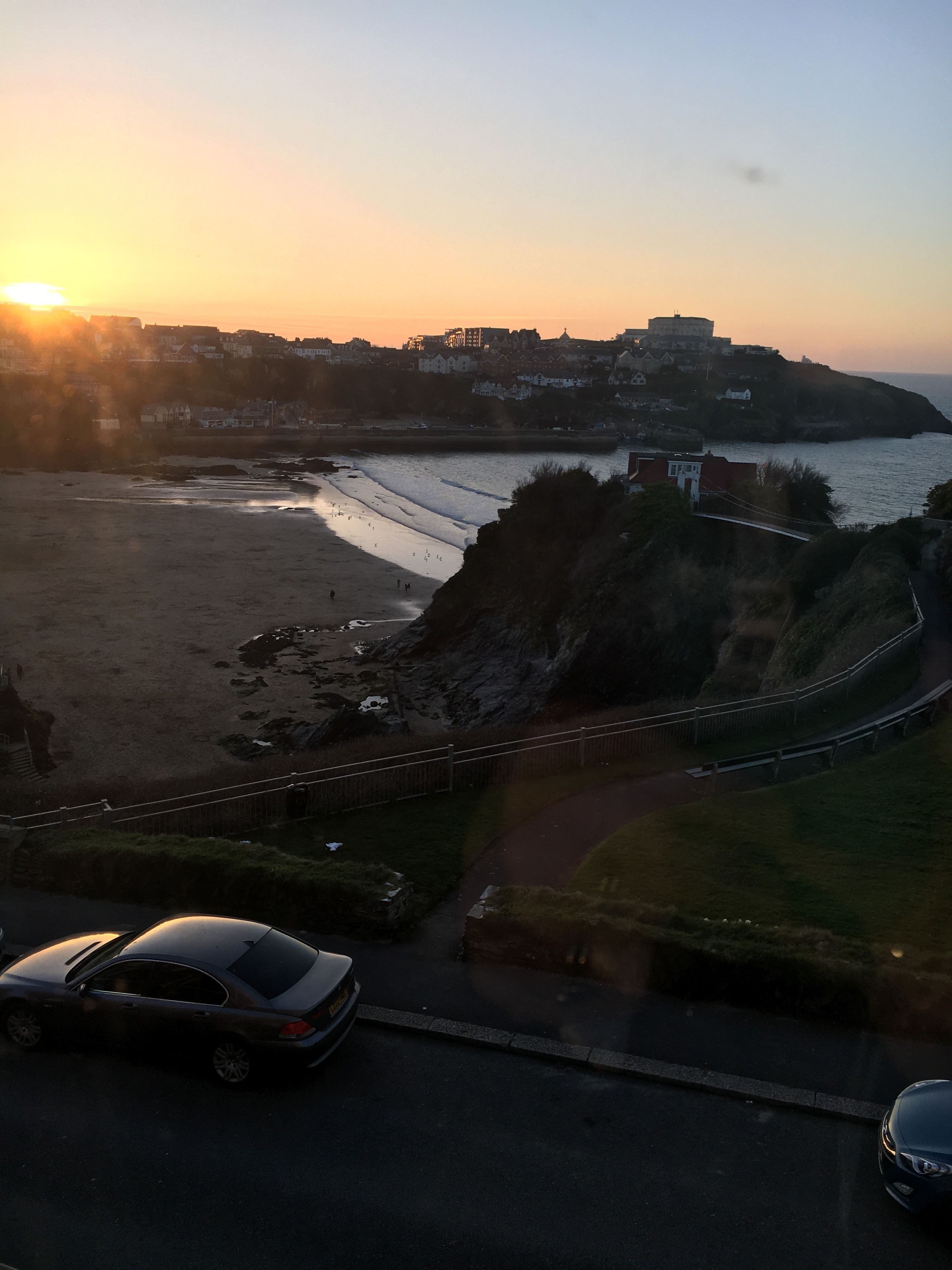 A coastal road with a car passing by, leading to a beach with a rocky cliff and a castle-like structure in the background.