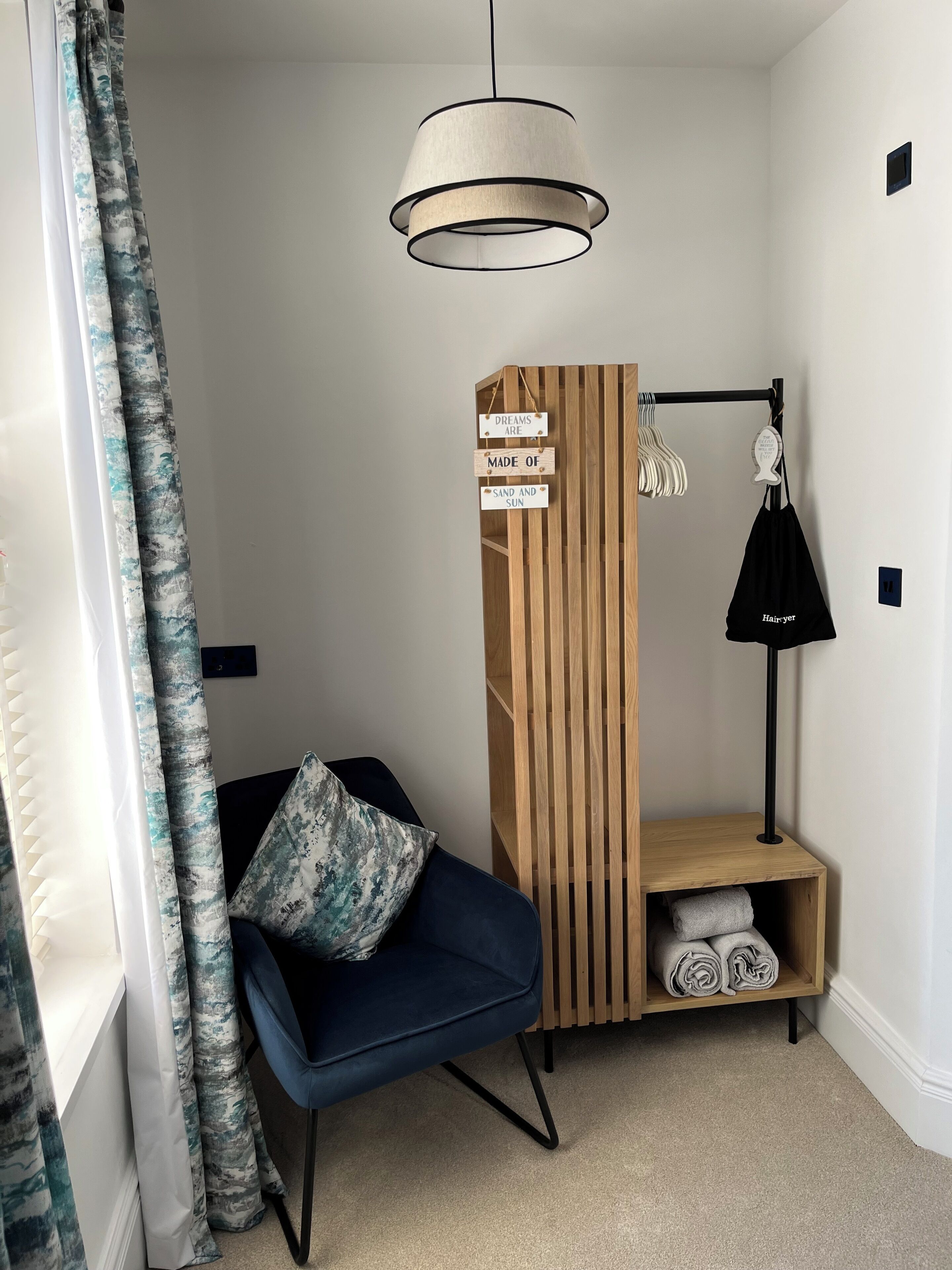 A corner room with a blue chair, a wooden shelf with towels, a hanging lamp, and a wall-mounted TV.