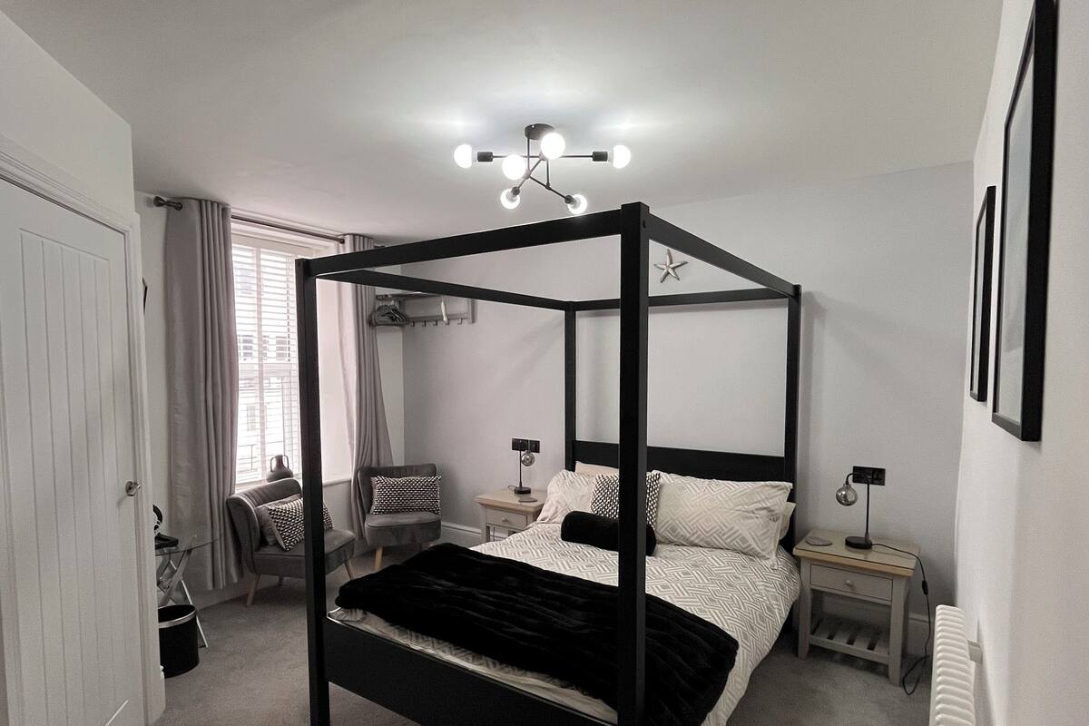 Una cama con dosel de estructura negra y ropa de cama blanca, una mesita de noche con lámpara, y un radiador en la pared.