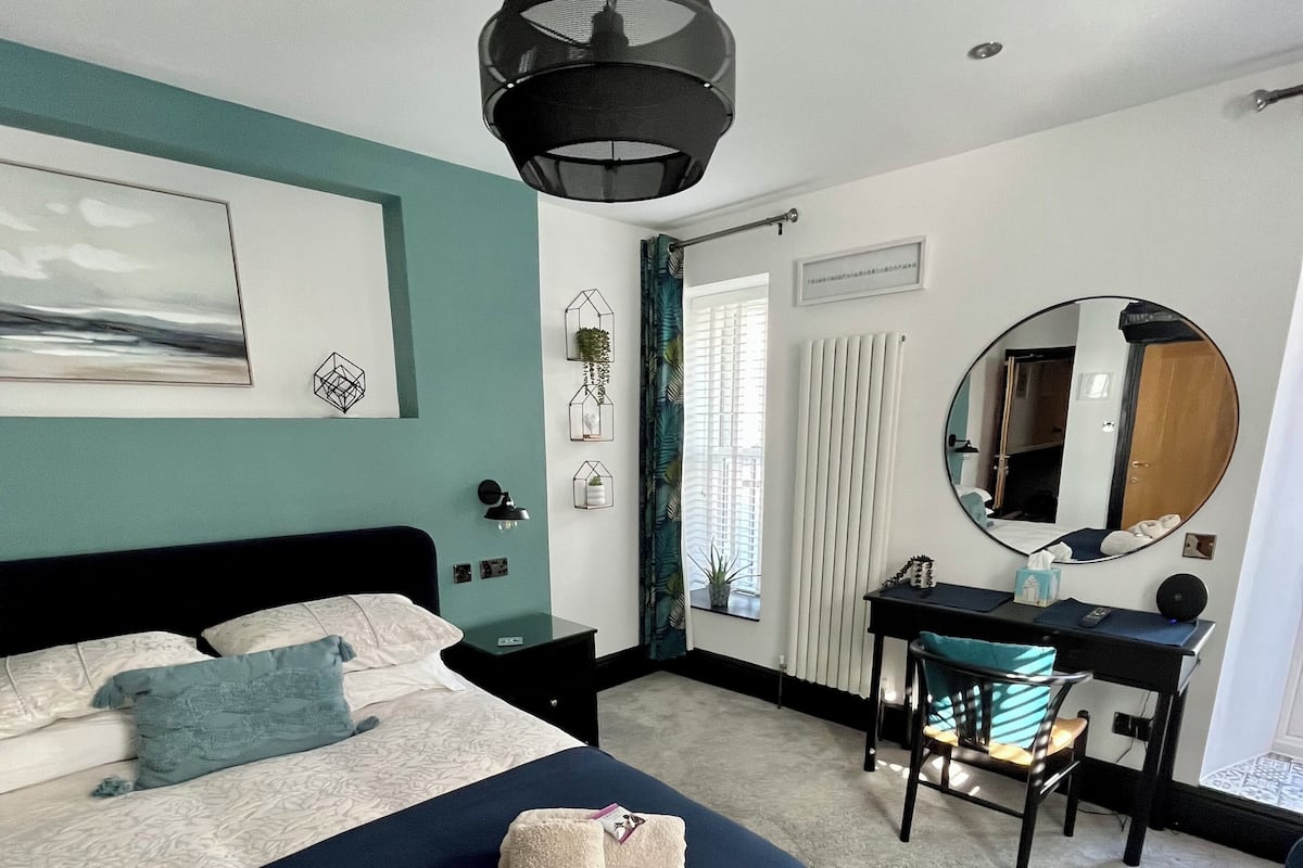 Un dormitorio con cama, escritorio, silla, espejo redondo y lámpara colgante.
