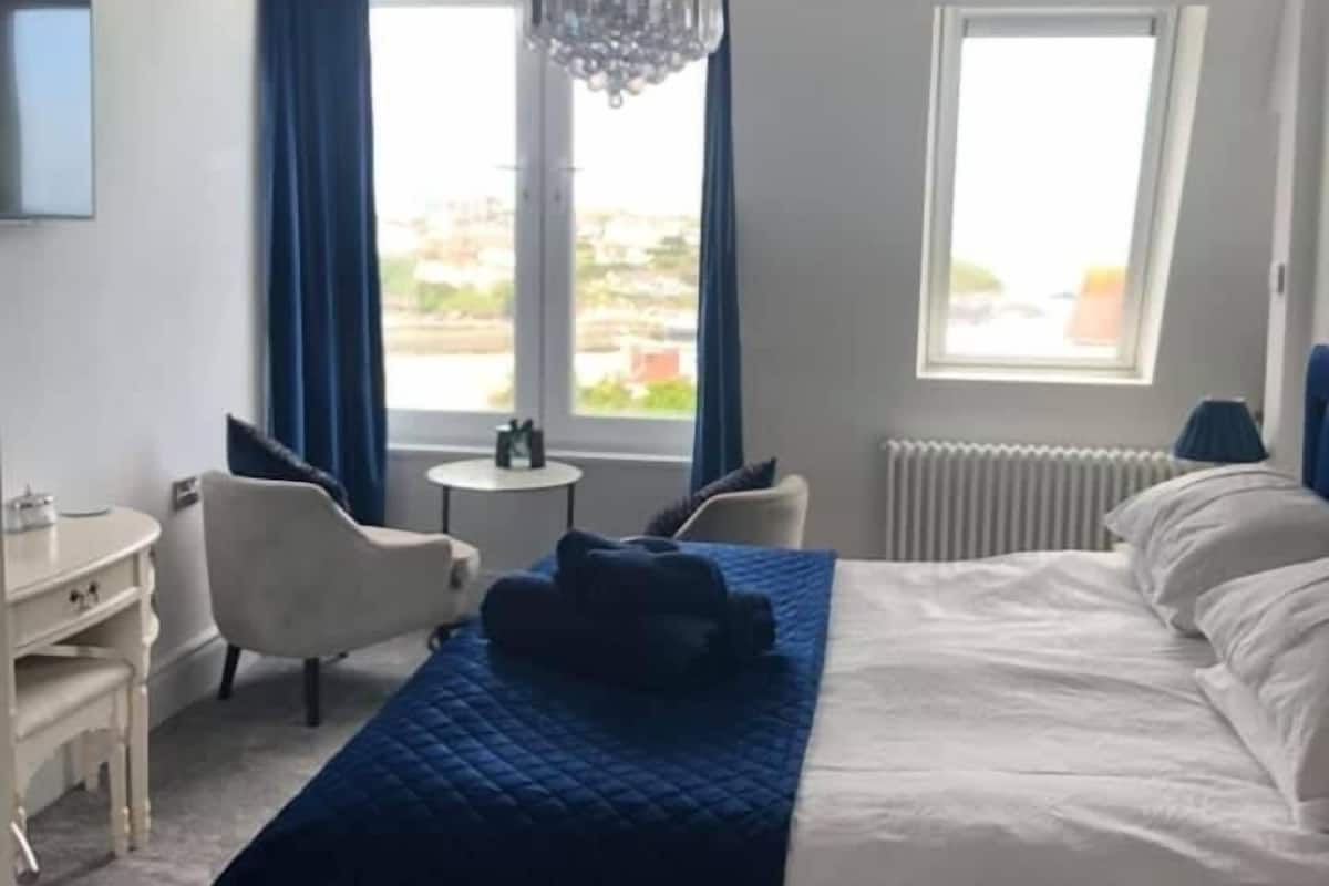 Habitación de hotel con cama, escritorio, silla, mesita, candelabro y ventana con cortinas.