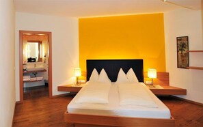 Une chambre d’hôtel avec un grand lit, deux tables de chevet, une commode, un miroir et une salle de bain visible à travers une porte ouverte.