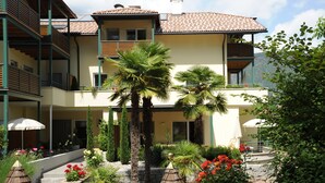 Un immeuble à plusieurs étages doté de balcons, un jardin avec des palmiers et des fleurs, et un sentier en pierre.