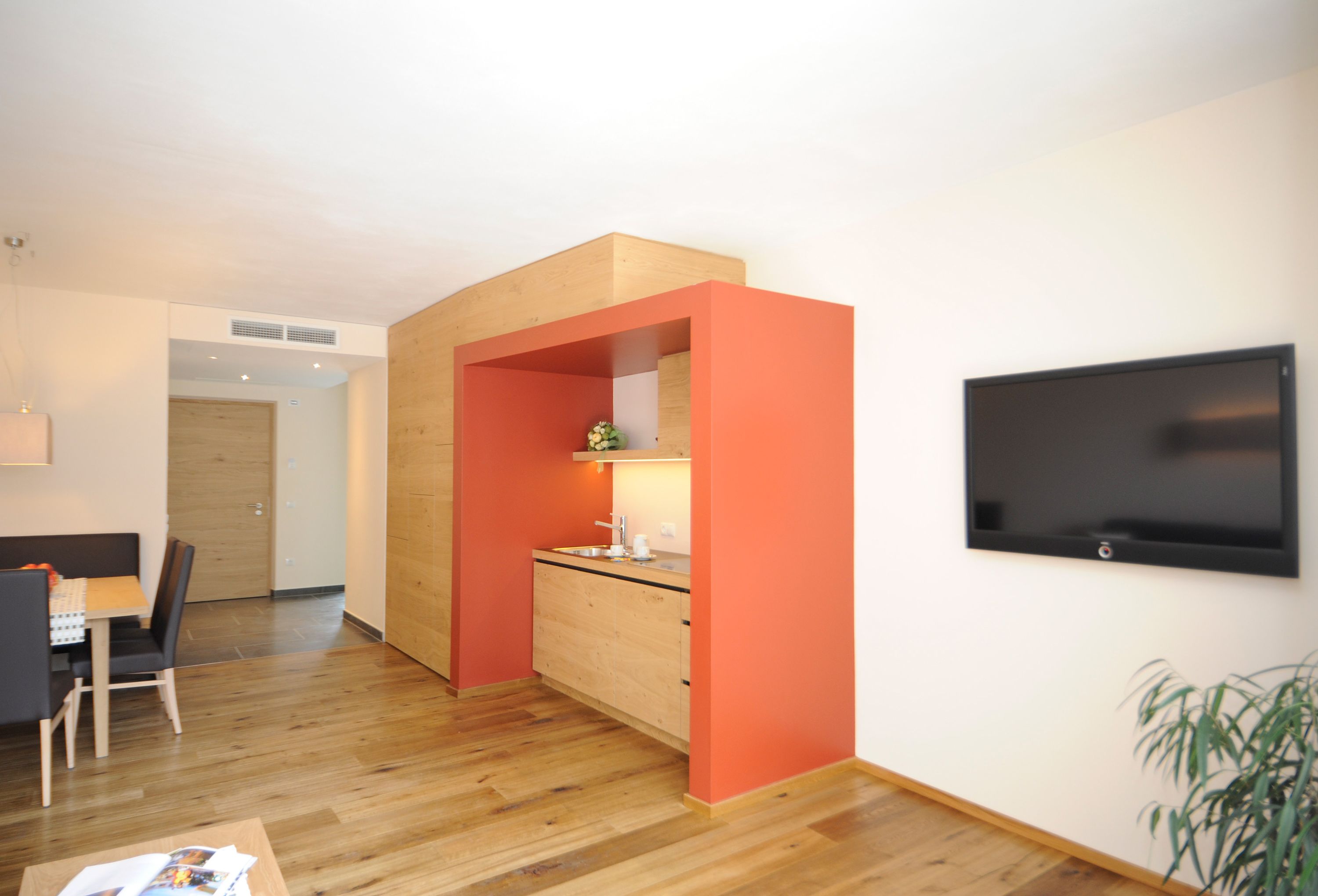 A modern kitchen with wooden cabinets and a built-in oven, a dining area with a table and chairs, and a flat-screen TV mounted on the wall.