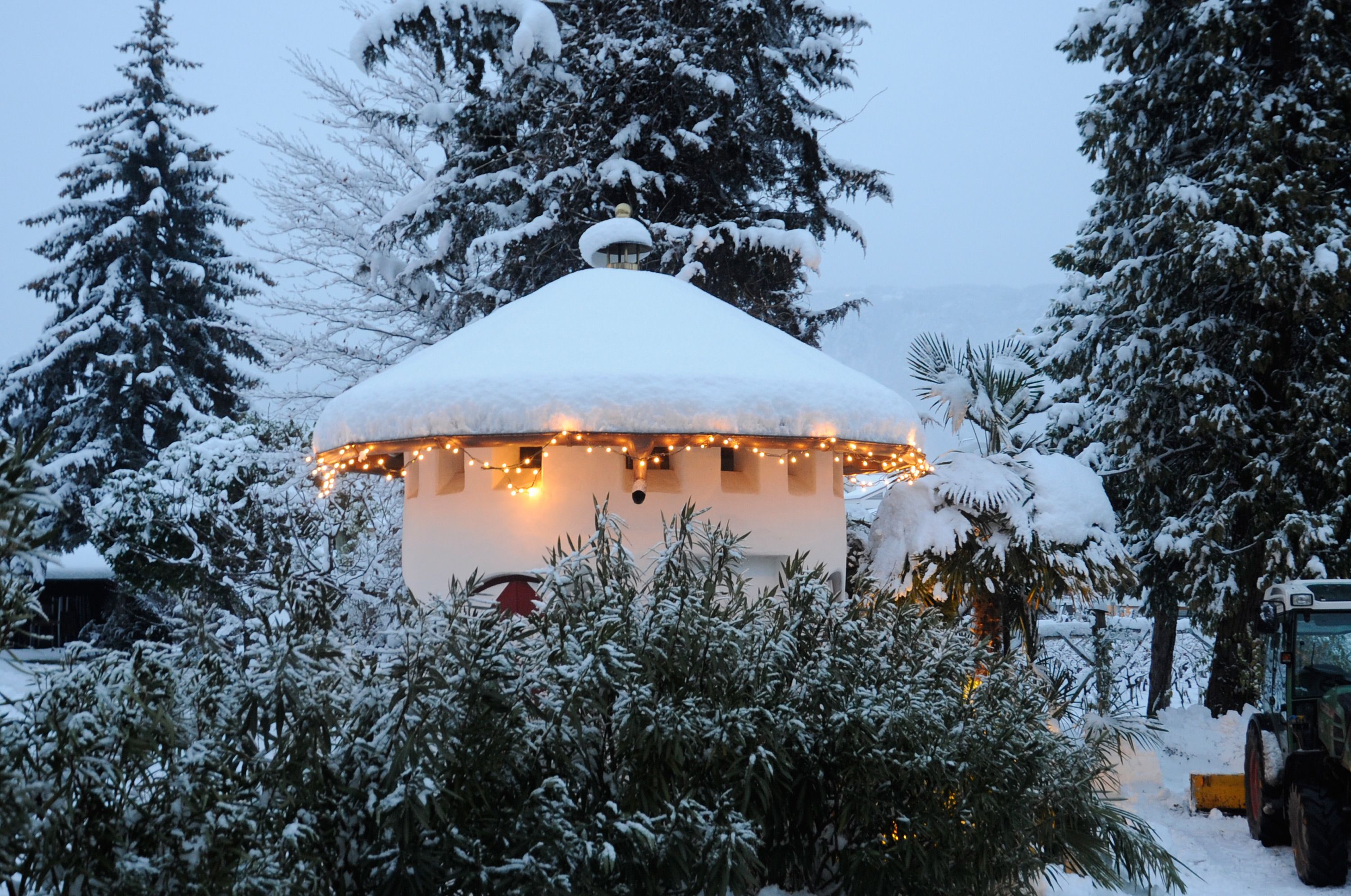 A snow-covered gazebo with string lights, surrounded by snow-laden trees.