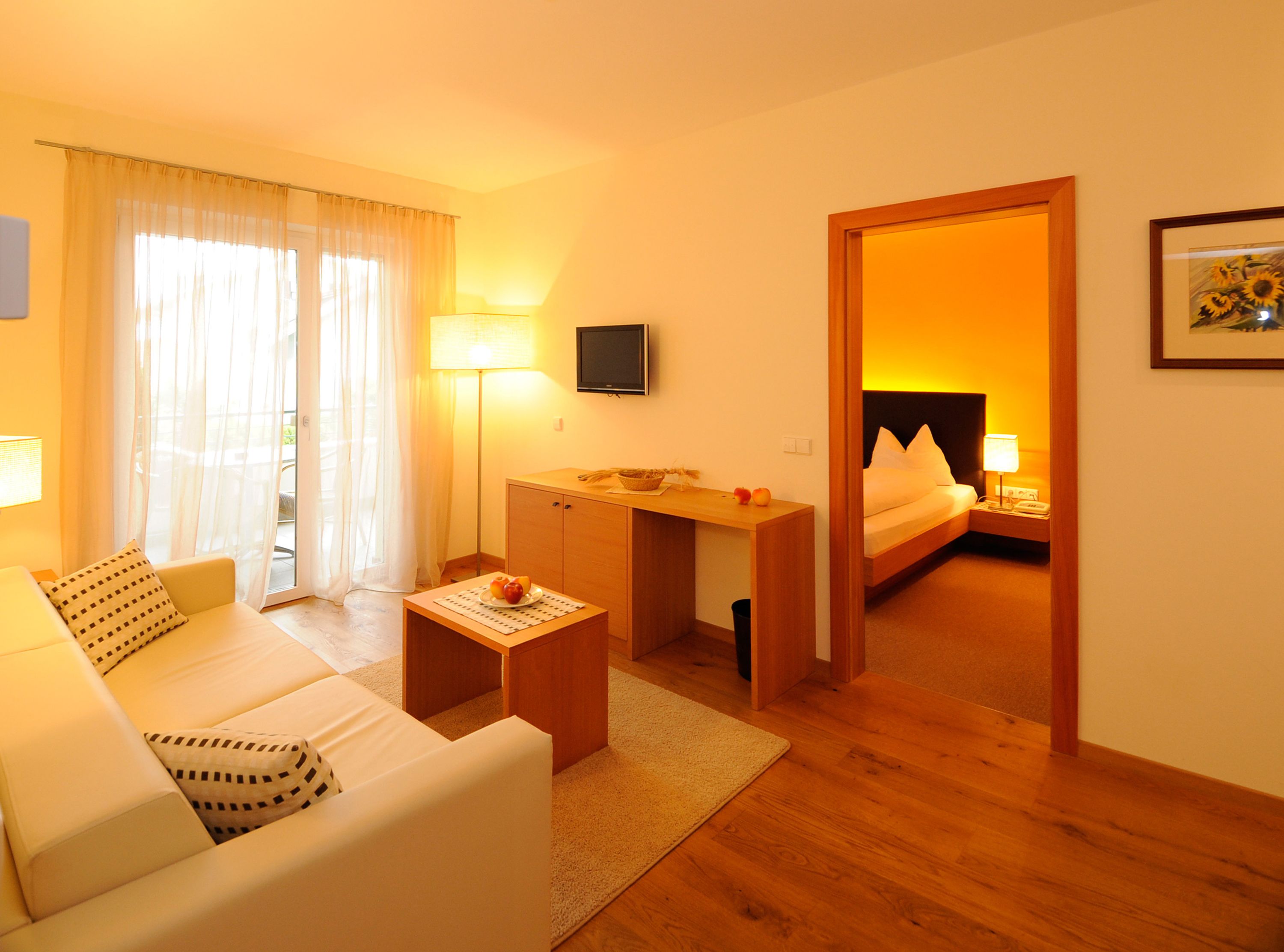 A hotel room with a sofa, a small table with fruit, a desk, a bed, and a TV on the wall.