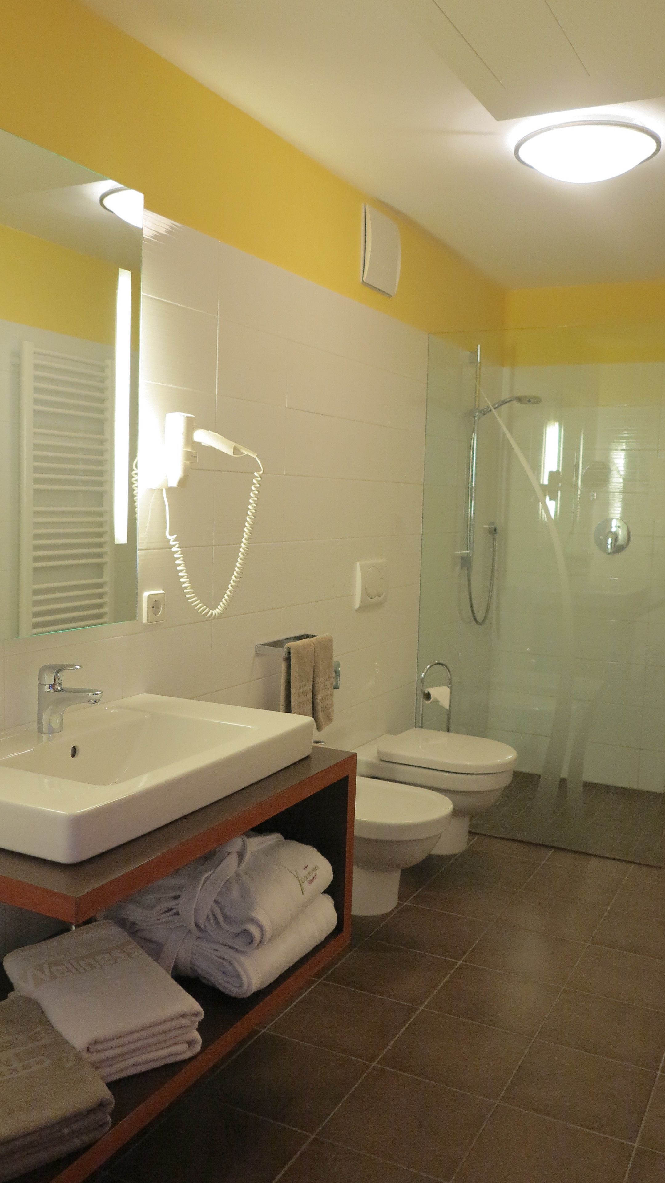 Une salle de bain moderne avec un lavabo, des toilettes et une douche, le tout de couleur blanche. On y trouve un porte-serviettes et une étagère avec des serviettes pliées.
