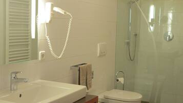 A modern bathroom with a white sink, toilet, and shower. There is a towel rack and a shelf with folded towels.