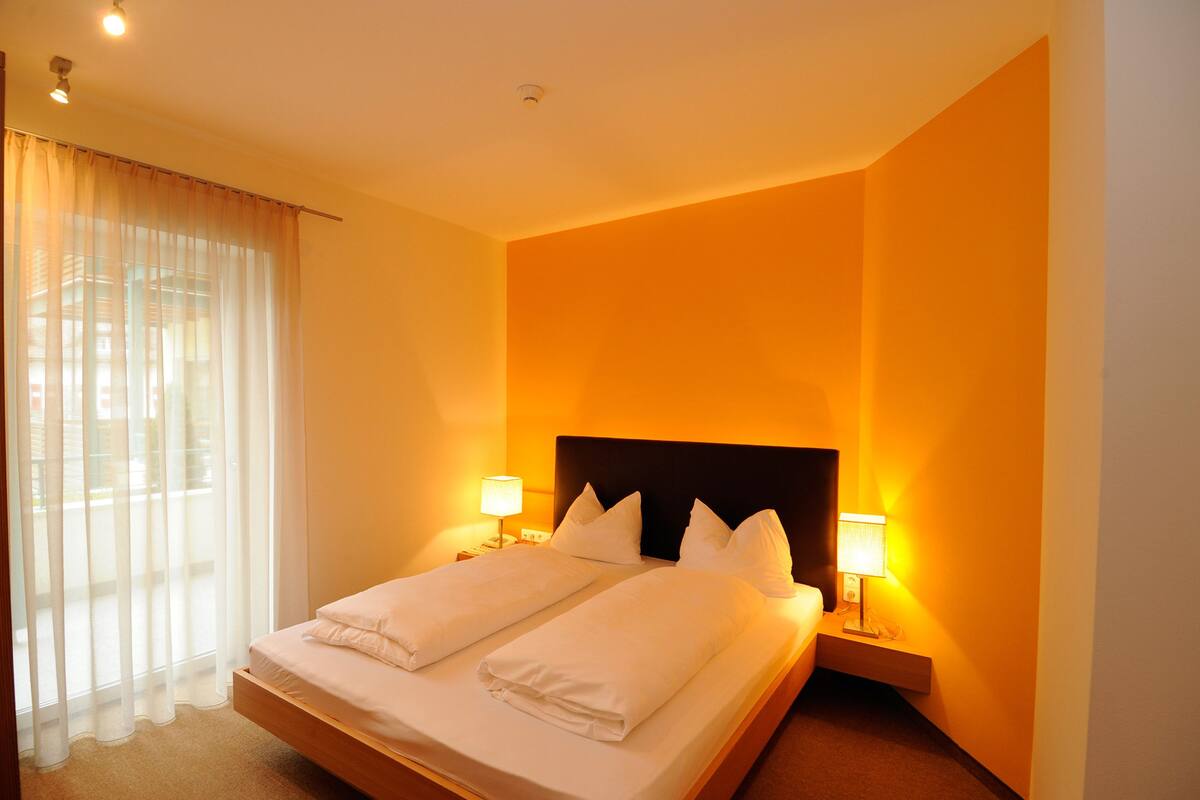 Ein Hotelzimmer mit einem großen Bett, zwei Nachttischlampen und einem Fenster mit transparenten Vorhängen.