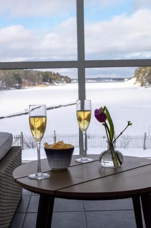 A table with two glasses of white wine, a bowl of snacks, and a vase with a single flower, set against a snowy landscape view. - Skeviks Gård (Värmdö)