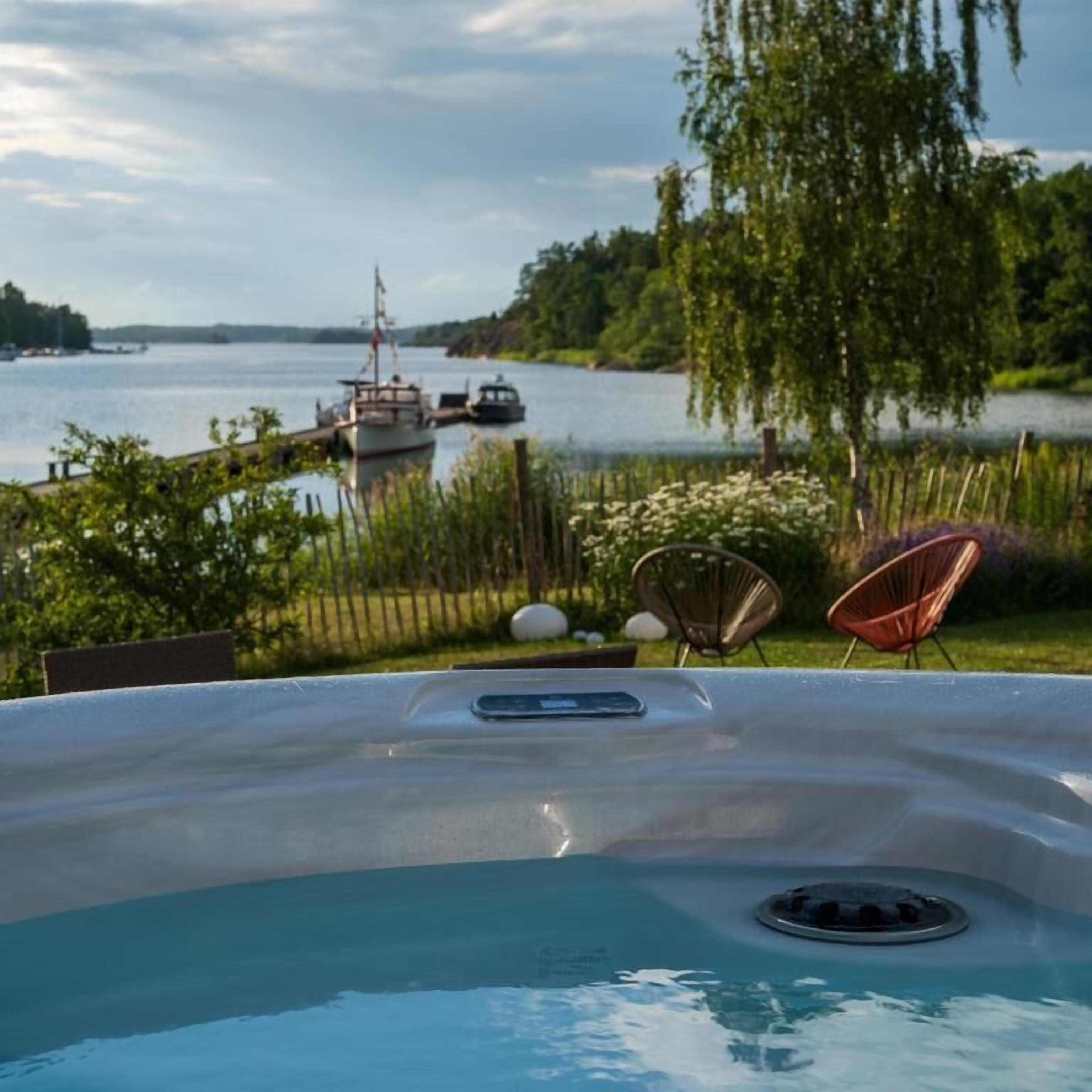 Ein Whirlpool mit Blick auf einen See und Boote, umgeben von Grünpflanzen und Gartenmöbeln.